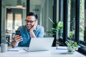 A man is smiling while using his phone and laptop